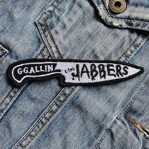 Puede incluir: Parche bordado con forma de cuchillo, con las palabras "G GALLIN" y "JABBERS" en letras blancas sobre un fondo blanco y negro. El parche está cosido sobre una chaqueta vaquera azul.