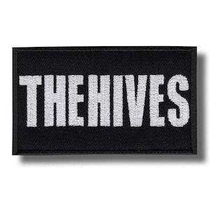 Può includere: Toppa rettangolare nera con la scritta bianca "THE HIVES". La toppa ha un bordo nero e un carattere bianco e in grassetto. La toppa è probabilmente per abbigliamento o accessori.
