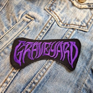Può includere: Toppa nera con la scritta "GRAVEYARD" ricamata in viola con un lettering stilizzato. La toppa ha una forma rettangolare curva ed è posizionata su una giacca di jeans.