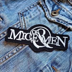 以下が含まれることがあります： ジーンズジャケットに縫い付けられた「MICE & MEN」の文字が入った白と黒の刺繍ワッペン。ワッペンは長方形で、上部と下部がカーブしています。テキストは太字の様式化されたフォントです。