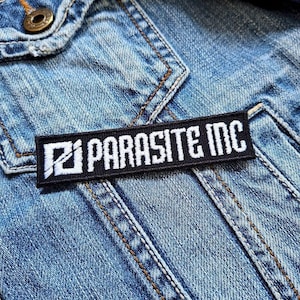 以下が含まれることがあります： 「PARASITE INC」と白文字で書かれた黒い長方形のパッチが、デニムジャケットに縫い付けられています。パッチの左側には白いグラフィックがあります。ジャケットは、ステッチが見えるライトブルーのデニムです。