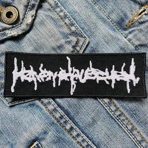Op de afbeelding: Een rechthoekige zwarte patch met witte geborduurde tekst. De witte tekst is een gestileerde bandnaam of logo in een gotisch lettertype. De patch is genaaid op een spijkerjasje.