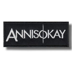 Könnte beinhalten: Ein rechteckiger schwarzer Aufnäher mit dem weißen gestickten Text "ANNISOKAY". Eine vertikale weiße Linie teilt das "O" in der Mitte des Wortes. Der Aufnäher hat eine strukturierte Optik und einen leichten Schatten.