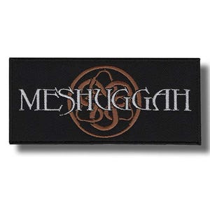Op de afbeelding: Een zwart rechthoekig embleem met het woord "MESHUGGAH" in wit, boven een bruin, gestileerd embleem. Dit embleem, waarschijnlijk voor kleding, toont het logo van een band.
