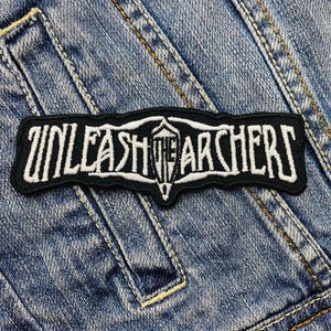 May include: A black and white embroidered patch with the text "UNLEASH THE ARCHERS" in a stylized font. The patch has a unique shape and is set against a denim background.