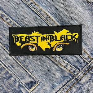 Puede incluir: Parche rectangular negro con el texto "BEAST IN BLACK" en amarillo, un gráfico amarillo y dos ojos amarillos estilizados. El parche está cosido a una chaqueta vaquera.