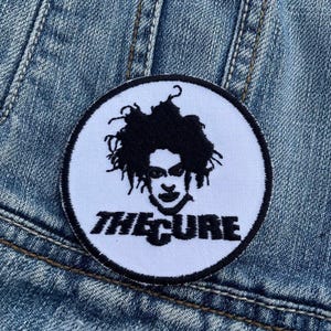 May include: A circular patch featuring a black and white graphic of a person with curly hair and the text "THE CURE" in black. The patch is sewn onto a blue denim jacket.