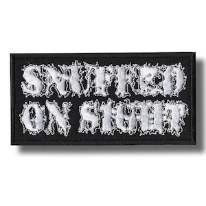 May include: A black rectangular patch with white embroidered text. The text reads "SNIFFED ON SHIT". The patch has a black border and a white shadow effect on the text.