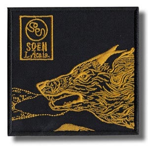 Puede incluir: Parche cuadrado negro con diseño de cabeza de lobo bordada en oro. El parche presenta el texto "SOEN Lakala" en un recuadro dorado. La boca abierta del lobo emite un diseño dorado estilizado.