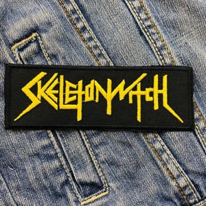 Puede incluir: Un parche rectangular negro con el texto "SKELETONWITCH" en amarillo, con letras estilizadas. El parche está sobre un fondo de mezclilla azul, con costuras y bolsillos visibles.
