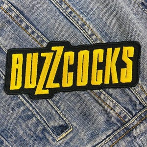 May include: A black and yellow embroidered patch with the text "BUZZCOCKS" in a bold, stylized font. The patch is rectangular with rounded corners and is set against a denim background.