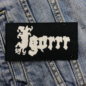 May include: A black rectangular patch with the white word "Igorr" in a stylized font. The patch is set against a denim jacket background. The lettering has a gothic or metal aesthetic.