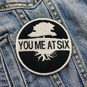 Può includere: Toppa rotonda ricamata in bianco e nero con la scritta bianca "YOU ME AT SIX". La toppa presenta un disegno di albero bianco sopra e sotto il testo, su una giacca di jeans.