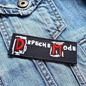 May include: A black rectangular patch with the words "Depeche Mode" in white and red lettering. The patch is sewn onto a blue denim jacket. The patch features a stylized "D" and "M" in red.