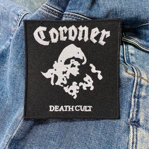 Könnte beinhalten: Schwarzer quadratischer Aufnäher mit weiß gesticktem Text und Grafik. Das Wort "CORONER" ist oben gewölbt, darunter ein schädelartiges Design. Der Text "DEATH CULT" befindet sich unten. Der Aufnäher befindet sich auf einer Jeansjacke.