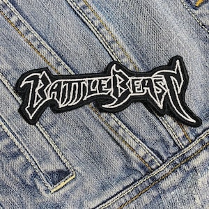 May include: Black and white embroidered patch with the text "BATTLE BEAST" in a stylized font. The patch is rectangular with a black background and white stitching. The patch is sewn onto a denim jacket.