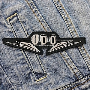 Op de afbeelding: Zwart en wit geborduurde patch met het woord "UDO" in een gestileerd lettertype. De patch heeft een rechthoekige vorm met puntige uiteinden en is op een spijkerjas genaaid.
