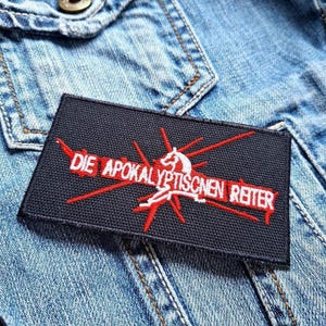 Puede incluir: Un parche rectangular negro con el texto "DIE APOKALYPTISCHEN REITER" en blanco y rojo. El parche presenta una imagen estilizada de un jinete y líneas rojas que irradian hacia afuera. El parche está sobre una chaqueta vaquera.