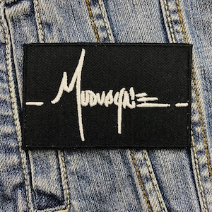 May include: A black rectangular patch with white script that reads "Mudvayne" in a stylized font. The patch is set against a denim background. The patch has a horizontal line above and below the text.