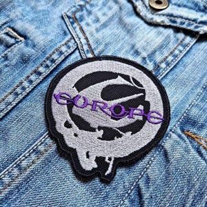 Op de afbeelding: Ronde geborduurde patch met het woord "COROPE" in paars. De patch heeft een zwart-wit ontwerp en is bevestigd op een spijkerjasje.