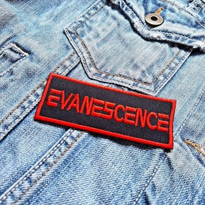 Op de afbeelding: Een rechthoekige zwarte patch met het woord "EVANESCENCE" in rode letters, omlijnd door een rode rand. De patch is bevestigd op een lichtblauwe spijkerjas, met zichtbare stiksels en een zakdetail.