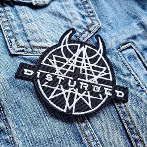 May include: A black and white embroidered patch with the word "DISTURBED" and a geometric design. The patch is circular with a banner at the bottom, set against a blue denim jacket.