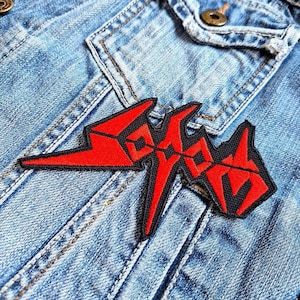 Pode incluir: Um patch bordado vermelho e preto com a palavra "Sodom" em uma fonte estilizada, costurado em uma jaqueta jeans azul claro. O patch tem uma borda preta e está posicionado na frente da jaqueta.