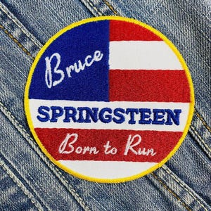 May include: A round embroidered patch with a yellow border, featuring a blue and red design. The patch includes the name "Bruce" in white script, the name "SPRINGSTEEN" in blue block letters, and the phrase "Born to Run" in white script.