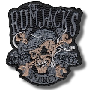 Może przedstawiać: Haftowana naszywka z czaszką w pirackim kapeluszu, skrzyżowanymi pistoletami i napisem "THE RUMJACKS" u góry i "SYDNEY" u dołu. Naszywka jest głównie czarna, z szarymi, beżowymi i białymi akcentami.