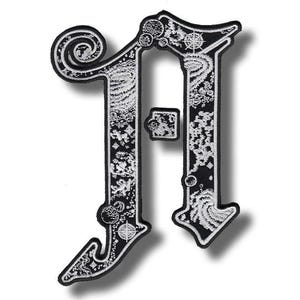 May include: Black and white embroidered patch in the shape of the letter "A". The patch features intricate designs with celestial and swirling patterns. The border is black, and the details are in white.