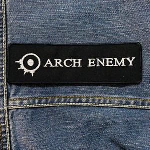 May include: Black rectangular patch with the text "ARCH ENEMY" in white, along with a white circular logo. The patch is sewn onto a blue denim fabric, with visible stitching and a pocket seam.