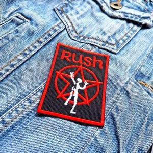 May include: A rectangular patch with the word "Rush" in red, above a red star and a white figure. The patch is on a blue denim jacket. The patch has a black background and a red border.
