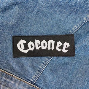 Peut inclure: Un écusson rectangulaire noir avec le mot "Coroner" en lettres gothiques blanches. L'écusson est cousu sur une veste en jean bleue, avec des coutures et des coutures visibles.