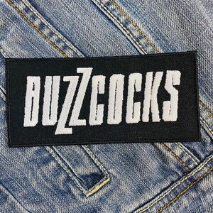 Könnte beinhalten: Ein schwarzer rechteckiger Aufnäher mit dem weißen Text "BUZZCOCKS" aufgestickt. Der Aufnäher befindet sich auf einem blauen Jeans-Hintergrund mit sichtbaren Nähten und Nähten. Der Text ist in einer fetten, stilisierten Schriftart.