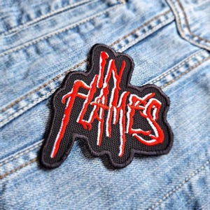 Könnte beinhalten: Ein schwarzer Aufnäher mit dem Schriftzug "IN FLAMES" in roter und weißer Schrift, auf eine hellblaue Jeansjacke genäht. Der Aufnäher hat einen gezackten, unregelmäßigen Rand.