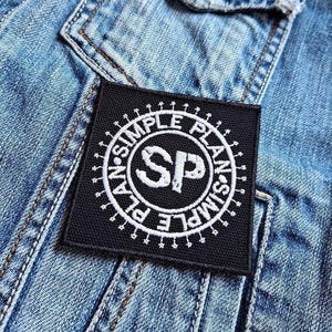 Puede incluir: Un parche cuadrado negro con las palabras "SIMPLE PLAN" en un diseño circular, rodeando las letras "SP" en blanco. El parche está cosido sobre una chaqueta vaquera azul claro.