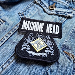 Puede incluir: Dos parches negros bordados sobre una chaqueta vaquera. El parche superior dice "MACHINE HEAD" en letras blancas. El parche inferior presenta un diseño de escudo con un diamante amarillo y detalles en gris.