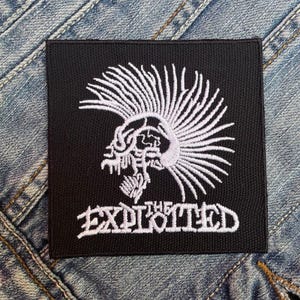 Può includere: Toppa quadrata nera con un teschio ricamato bianco con cresta e la scritta "THE EXPLOITED". La toppa, pensata per l'abbigliamento, è presentata su uno sfondo in denim, suggerendo il suo utilizzo come accessorio.