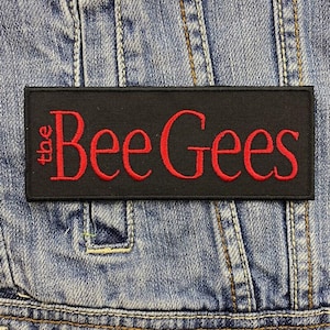 May include: Black rectangular patch with the embroidered text "the Bee Gees" in red. The patch is affixed to a denim jacket. The text is in a cursive font.