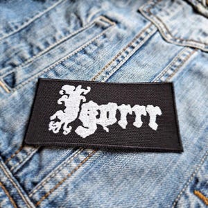 May include: A black rectangular patch with white embroidered text that reads "Gorr". The patch is placed on a light blue denim jacket. The text has a stylized, gothic font.