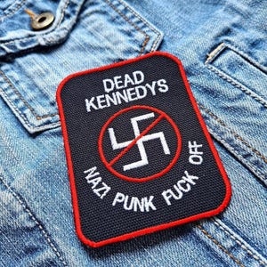 Könnte beinhalten: Ein rechteckiger schwarzer Aufnäher mit rotem Rand, der den Text "DEAD KENNEDYS" über einem weißen Hakenkreuzsymbol mit einem roten Strich durchgestrichen zeigt. Darunter steht "NAZI PUNK FUCK OFF" in weißer Schrift. Der Aufnäher befindet sich auf einer blauen Jeansjacke.