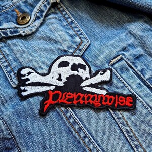 Op de afbeelding: Geborduurde patch met een wit schedel- en kruisbottenontwerp op een zwarte achtergrond. Het woord "Dreamwise" is in het rood gestikt. De patch is bevestigd op een spijkerjasje.