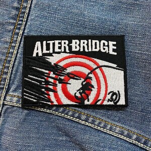 Op de afbeelding: Een rechthoekige zwarte patch met de witte tekst "ALTER BRIDGE". De patch heeft een rood-wit doelontwerp met een zwart silhouet. De patch is genaaid op een blauwe denim stof.