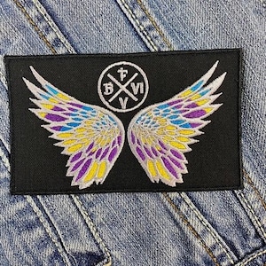 Könnte beinhalten: Schwarzer rechteckiger Aufnäher mit gestickten Engelsflügeln in Gelb, Lila und Blau. Ein kreisförmiges Emblem mit den Buchstaben B, V und VI ist über den Flügeln zentriert. Der Aufnäher ist vor einem Jeanshintergrund platziert.