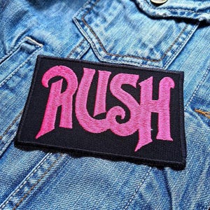 May include: A black rectangular patch with the word "RUSH" embroidered in pink, set against a blue denim jacket. The patch features a stylized font, and the denim jacket shows visible stitching and a pocket.