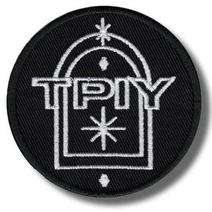 May include: A round, black embroidered patch with white stitching. The design features the letters "TPY" within a stylized architectural shape, with starburst and dot accents. The patch has a clean, modern aesthetic.