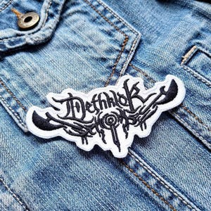 Puede incluir: Un parche blanco con texto y diseño bordado en negro, que dice "Dethklok". El parche tiene una forma irregular con detalles en negro y está cosido a una chaqueta vaquera. El parche es un artículo relacionado con la música.
