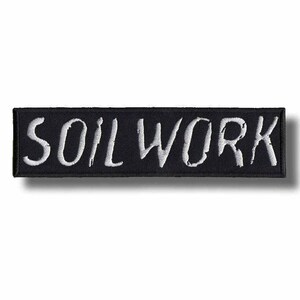 May include: A black rectangular patch with the word "SOIL WORK" embroidered in white. The patch has a black border and is likely designed to be sewn or ironed onto clothing or other fabric items.