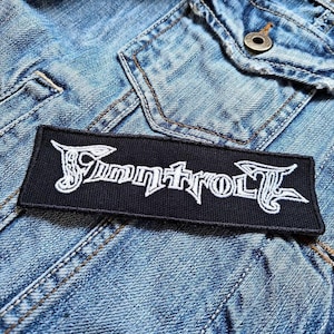 Puede incluir: Un parche rectangular negro con la palabra "Tummytroll" bordada en blanco, con letras góticas estilizadas. El parche está colocado sobre una chaqueta vaquera azul claro. El parche es rectangular.