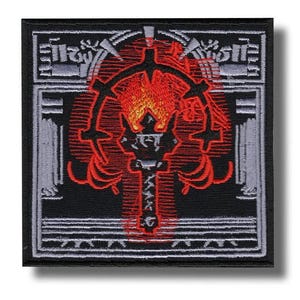 May include: Square black patch with a red and orange emblem. The emblem features a central cross-like shape with a flame design at the top and a circular design around it. The patch has a gray border with a detailed pattern.